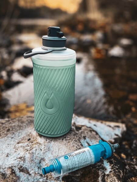 Składana butelka Flux™ HydraPak<sup>®</sup> z zamontowanym filtrem do wody HydraPak<sup>®</sup> (28 mm) położona na kamieniu przy strumieniu. Foto: Rigad