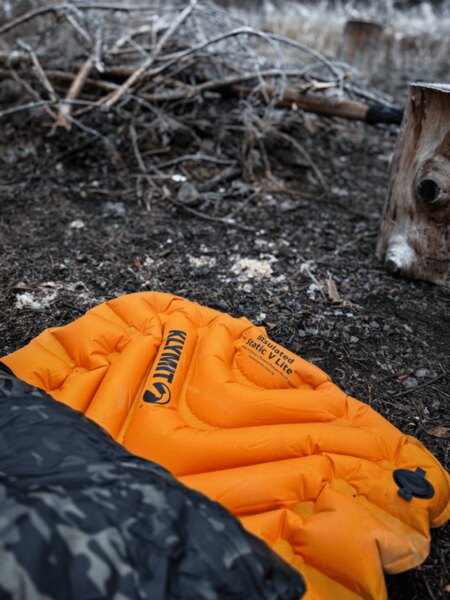 Nadmuchiwany materac Klymit Insulated Static V Lite w pomarańczowym kolorze na ziemi przy ognisku, częściowo zakryty śpiworem z wzorem maskującym. Izolacja od zimna i wilgoci podczas spania w terenie. Foto: Rigad